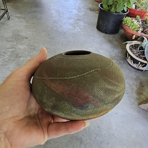 May include: A round, low-profile ceramic vase with a textured, matte surface in shades of olive green, brown, and hints of reddish-orange. The vessel features a small, circular opening at the top and a subtle, raised horizontal line detail.
