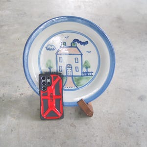 Könnte beinhalten: Eine handbemalte Keramikplatte mit einer Hausszene, mit blauem Rand und Akzenten. Neben dem Teller befindet sich eine rote und schwarze Handyhülle. Der Teller wird auf einem Holzständer präsentiert.