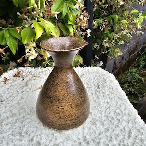 May include: A ceramic vase with a wide, flared rim and a rounded, bulbous body. The surface features a mottled, speckled glaze in shades of dark brown, tan, and black, creating a textured appearance across the entire exterior of the vessel.