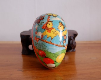 May include: Vintage Easter egg-shaped cardboard container with a colorful illustration of two chicks on a branch, surrounded by flowers and eggs. The container is blue, green, yellow, red, and white.