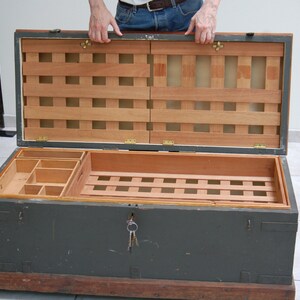 May include: A vintage wooden military chest with a green painted exterior and a wood interior. The chest has a hinged lid and a keyhole with keys in the lock. The interior has a small compartment with dividers and a larger compartment with a slatted bottom.