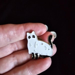 May include: An enamel pin shaped like a white cat with black ears, black paws, and a black tail. The cat has black eyes and small black markings on its body, including a cross shape on its back. The pin features a gold-toned metal outline.