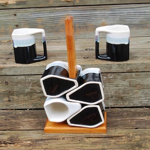 May include: Set of six black and white ceramic mugs with a wooden stand. The mugs have a white rim and a black glaze with a white stripe. The stand is made of wood and has a rectangular shape.