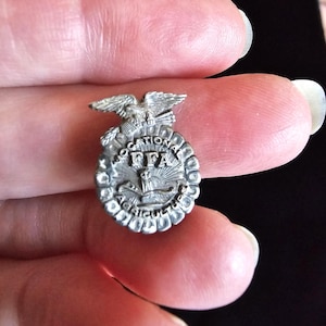 May include: A small silver-toned metal pin featuring an eagle perched atop a circular emblem. The emblem displays the text VOCATIONAL AGRICULTURE and the letters FFA, with an owl and a rising sun depicted in the center of the circular design.
