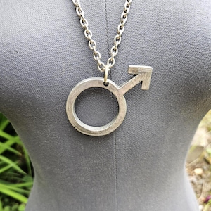 May include: A silver-toned metal pendant shaped like the male gender symbol, featuring a circular ring with an upward-pointing arrow. The pendant is suspended from a matching silver-toned metal chain link necklace with a textured, matte finish.