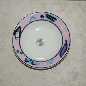 Puede incluir: Un pequeño cuenco de cerámica blanca con un borde rosa y diseños abstractos en negro, azul y amarillo. El cuenco está sobre una superficie beige claro.