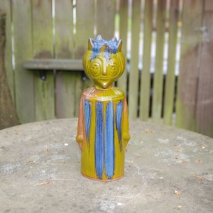 May include: A ceramic figurine of a king wearing a crown and a yellow and blue robe. The figurine is standing on a gray surface.