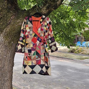 May include: A long-sleeved coat featuring a patchwork design composed of various geometric shapes in diverse colors and patterns. The garment has a bright orange inner lining, a wrap-style front closure, and a decorative circular button fastener.