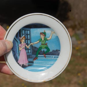May include: A small, white decorative plate with a gold rim. The plate features a colorful illustration of Peter Pan and Wendy from the classic Disney film. Peter Pan is wearing a green outfit, and Wendy is in a pink dress.