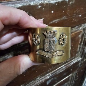 Puede incluir: Un servilletero de latón con detalles en relieve. El anillo presenta un escudo con una corona, un escudo y la palabra "BREST" en una pancarta. Dos elementos florales decorativos flanquean el escudo. El anillo está sostenido por una mano.