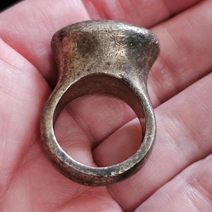 May include: A thick, heavy-looking ring with a weathered, dark metallic finish. The band is wide and solid, featuring a prominent, rounded top section that flares outward from the shank. The surface shows visible scratches and a dull, aged patina.
