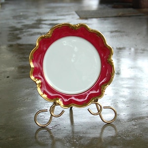 May include: A decorative plate with a red and gold design, featuring a white centre, displayed on a gold metal stand.
