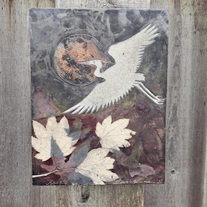 May include: A rectangular artwork featuring a white bird in flight against a backdrop of a full moon. Below the bird are three light-colored leaves. The artwork has a textured, multi-colored background with a wooden frame.