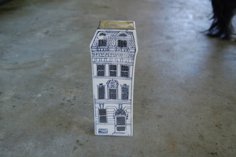 May include: A white ceramic figurine of a tall building with black details. The building has multiple windows and a decorative roof.