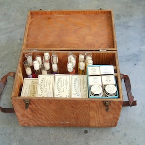 May include: Open wooden box with a hinged lid, filled with glass vials, small boxes, and round containers. The box has a leather handle and metal clasps. Several boxes are labeled "SANDSTEEL".