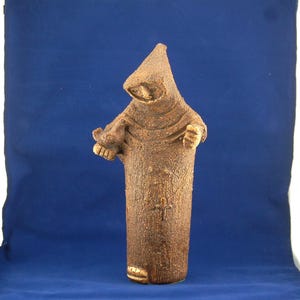 May include: A brown ceramic statue of a robed figure with a hood, holding an object in its hands. The statue has a textured surface and a cross design on the front. The figure is standing on a base.