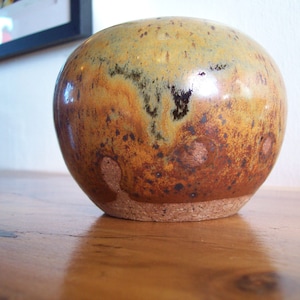 May include: A ceramic vase with a brown, yellow, and black glaze. The vase has a round shape and a textured surface.
