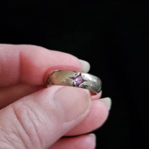 May include: A silver-toned metal band ring featuring a single, small, round, pink-colored stone set into the center of the band. The surface of the ring has a smooth, polished finish with some visible signs of wear and minor surface scratches.