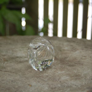 May include: A clear glass paperweight with a swirling, abstract design. The paperweight is sitting on a gray, textured surface.