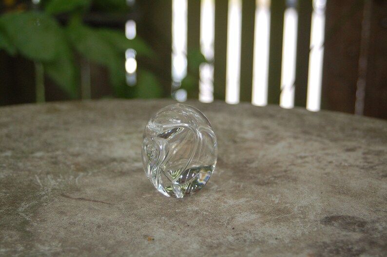 May include: A clear glass paperweight with a swirling, abstract design. The paperweight is sitting on a gray, textured surface.