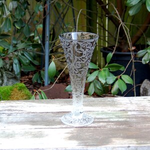 Puede incluir: Una flauta de champán de vidrio transparente con un diseño floral grabado. La flauta tiene un tallo alto y delgado y una copa ancha y redonda.