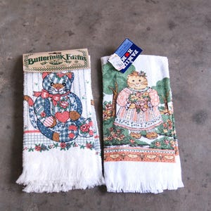 May include: Two white kitchen towels with decorative designs. One features a teddy bear in a plaid pattern with a red bow, the other a cat in a dress holding flowers. Both have fringe at the bottom and tags.