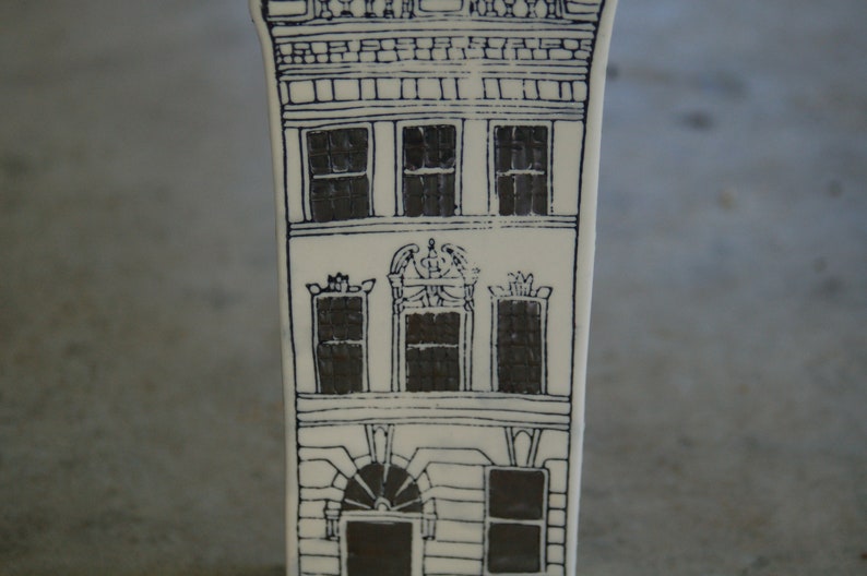 May include: A white ceramic building with black line drawings of windows and a decorative design above the windows. The building has a simple, minimalist style.