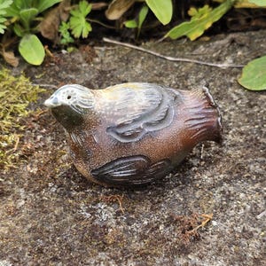 Może przedstawiać: Ceramiczna figurka ptaka w odcieniach brązu, beżu i szarości. Ptak ma szczegółowe wzory skrzydeł i piór, z lekko otwartym dziobem. Figurka spoczywa na teksturowanej, szarej kamiennej powierzchni, otoczonej zielenią.