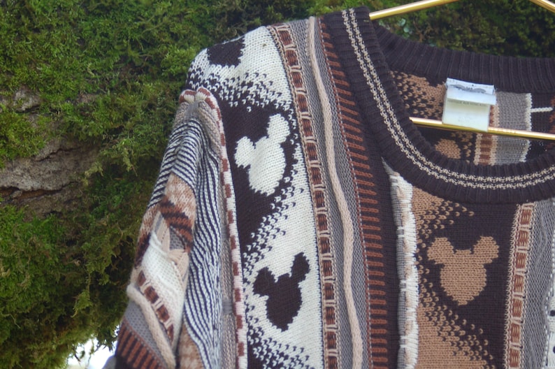 Peut inclure: Gros plan sur un pull en tricot marron, beige et blanc avec des silhouettes de Mickey Mouse. Le pull a un col rond et une vari&eacute;t&eacute; de motifs textur&eacute;s. Le v&ecirc;tement est suspendu &agrave; un cintre dor&eacute; sur fond de mousse verte.