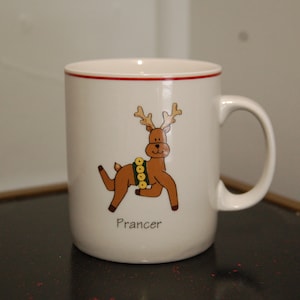 May include: White ceramic mug with a red rim and a cartoon reindeer with a green and yellow collar. The reindeer is brown and has a name "Prancer" printed below it.