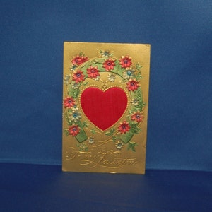 May include: A gold-coloured Valentine's Day card with a red heart in the centre of a horseshoe-shaped floral wreath. The card reads "To my Valentine".