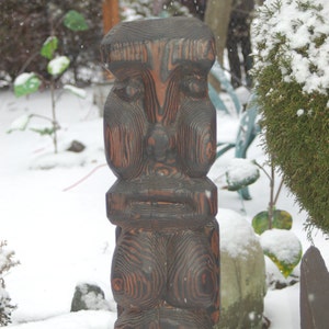 Pode incluir: Uma escultura de madeira marrom escura de um rosto humano com uma superfície texturizada. A escultura está em pé sobre uma superfície nevada.