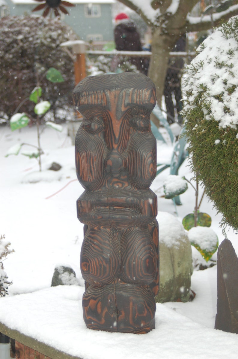 Op de afbeelding: Een donkerbruine houten sculptuur van een menselijk gezicht met een getextureerd oppervlak. De sculptuur staat op een besneeuwde ondergrond.