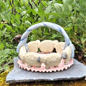 May include: A decorative sheep basket sculpture. The sculpture features six sheep around a pink base, with a blue and white handle. The sculpture is set on a gray stone, surrounded by greenery.