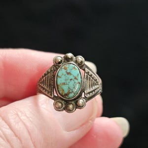 May include: A silver ring featuring an oval turquoise stone with brown matrix inclusions. The setting includes three small silver spheres above and below the stone, with triangular geometric engravings on the sides of the band.
