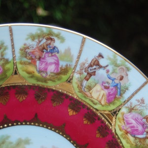 Peut inclure: Une assiette en porcelaine vintage avec un bord rouge et or. L'assiette pr&eacute;sente un motif bleu et blanc de couples dans un jardin.
