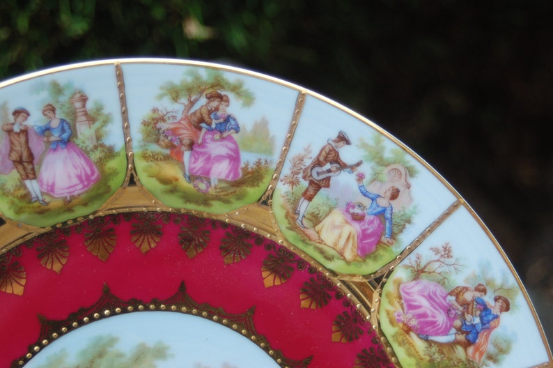 Peut inclure: Une assiette en porcelaine vintage avec un bord rouge et or. L'assiette pr&eacute;sente un motif bleu et blanc de couples dans un jardin.
