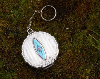 Op de afbeelding: Een zilveren compactpoederdoosje met een ketting en ringbevestiging. De compactpoederdoos heeft een geribbeld ontwerp met een centraal turquoise ovaal met een roze roos. De compactpoederdoos staat op een mosachtige achtergrond.