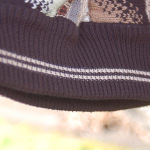 Peut inclure: Gros plan sur un bonnet en tricot marron fonc&eacute; avec un bord c&ocirc;tel&eacute;. Le bord comporte deux rayures blanches. Le haut du chapeau pr&eacute;sente un motif en nuances de marron, beige et blanc.