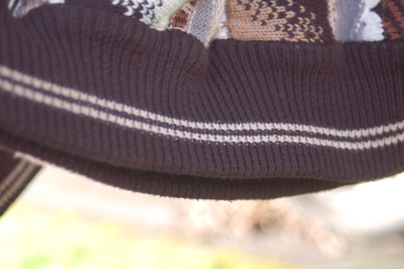 Peut inclure: Gros plan sur un bonnet en tricot marron fonc&eacute; avec un bord c&ocirc;tel&eacute;. Le bord comporte deux rayures blanches. Le haut du chapeau pr&eacute;sente un motif en nuances de marron, beige et blanc.