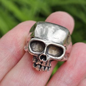 May include: A silver-toned metal ring featuring a detailed, three-dimensional skull design. The skull has deep, darkened eye sockets, a small nasal cavity, and visible teeth. The metal surface has a textured, slightly weathered, and matte finish.
