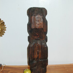 Op de afbeelding: Een donkerbruin houten beeld van een gestileerde menselijke figuur met een gegraveerd gezicht. Het beeld staat op een houten tafel met een groene appel ervoor.