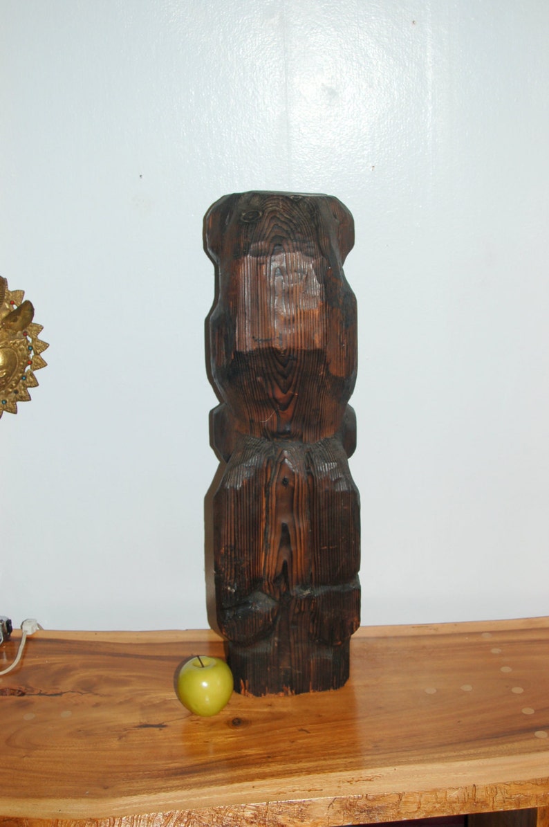 Op de afbeelding: Een donkerbruin houten beeld van een gestileerde menselijke figuur met een gegraveerd gezicht. Het beeld staat op een houten tafel met een groene appel ervoor.