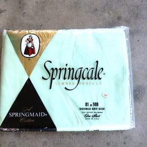 May include: A packaged Springcale double bed sheet in a pale green hue. The packaging displays the brand name, a logo with a woman in a red dress, and the dimensions 206 x 274 cm. The sheet is made of cotton.