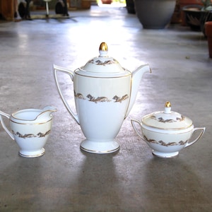 May include: A vintage white porcelain tea set with gold trim and decorative accents. The set includes a teapot, creamer, and covered sugar bowl. The teapot has a tall, slender shape with a curved handle. The creamer and sugar bowl have matching designs.