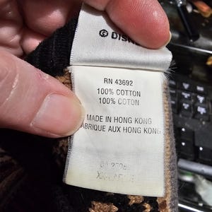 Peut inclure: Gros plan sur une &eacute;tiquette avec le logo Disney, RN 43692, et du texte indiquant 100% coton. L'&eacute;tiquette indique &eacute;galement "Made in Hong Kong" et "ABRIQUE AUX HONG KONG". L'&eacute;tiquette est tenue par une personne.