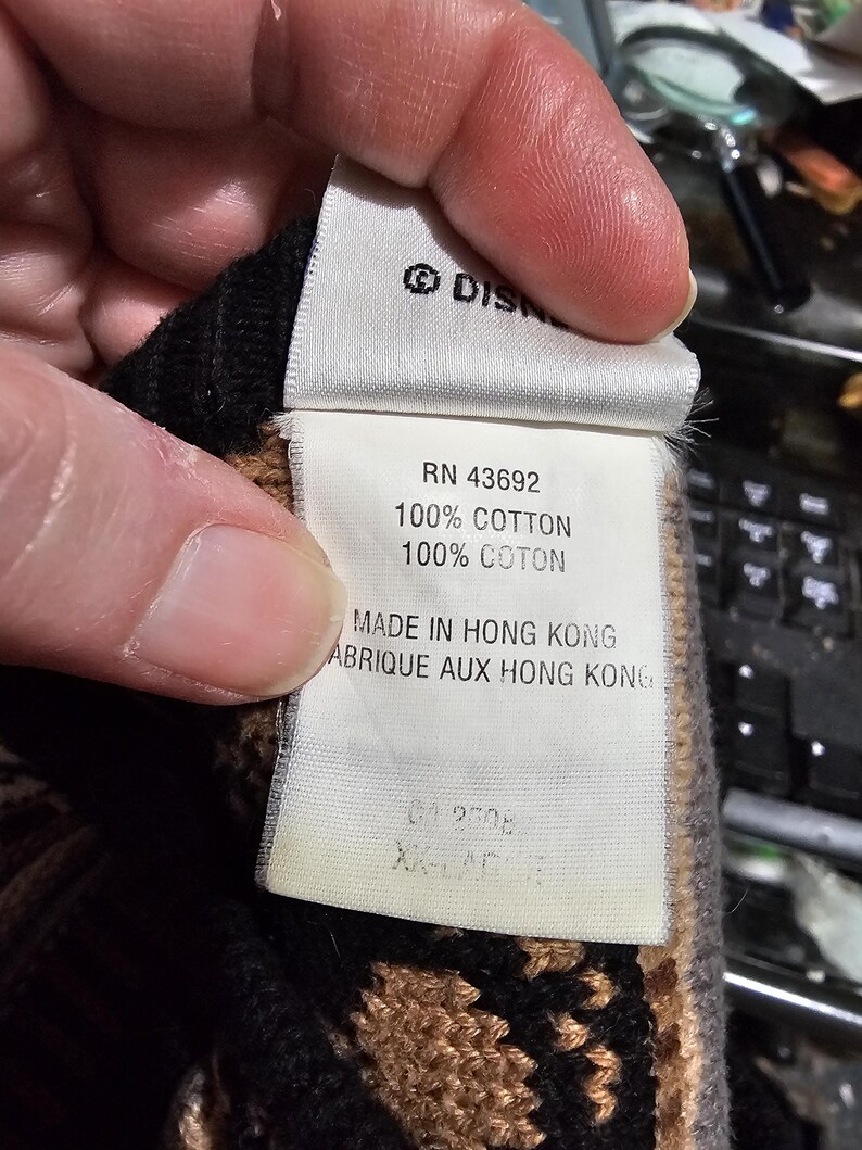 Peut inclure: Gros plan sur une &eacute;tiquette avec le logo Disney, RN 43692, et du texte indiquant 100% coton. L'&eacute;tiquette indique &eacute;galement "Made in Hong Kong" et "ABRIQUE AUX HONG KONG". L'&eacute;tiquette est tenue par une personne.