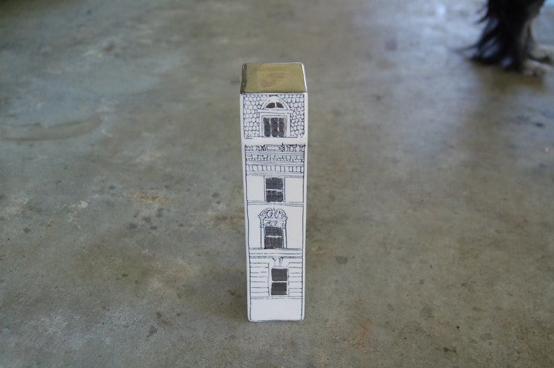 May include: A white ceramic building with a gold top, featuring three windows and a balcony. The building is drawn in black ink.