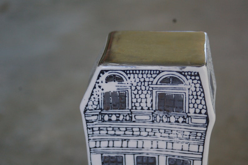 May include: A white ceramic house figurine with black and white details. The roof is a flat, silver color. The house has two windows with black shutters and a brick facade.