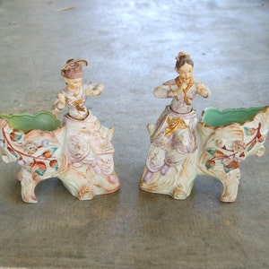 May include: Pair of antique porcelain planters, each featuring a figure in traditional attire. The planters are cream-colored with floral and leaf accents in pink, green, and gold. The interior of each planter is a light green.